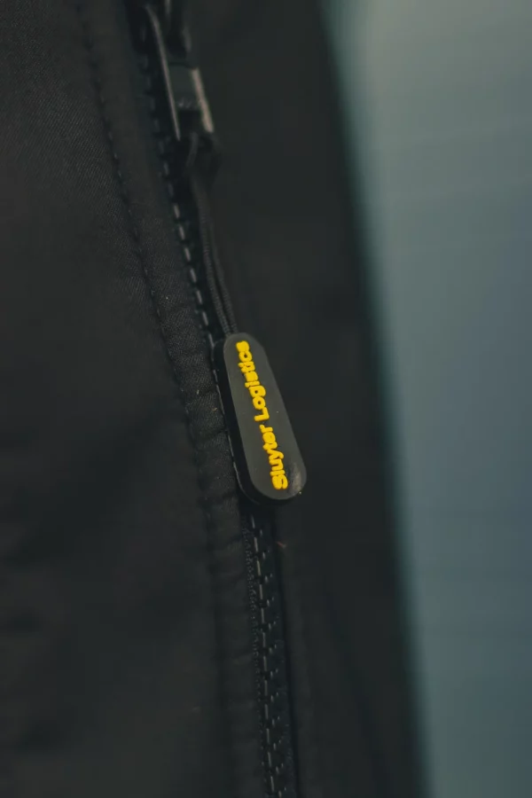 Detail van een zwarte werkjas met geel Sluyter Logistics-label aan de rits