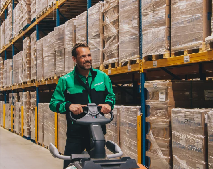 Magazijnmedewerker in groene werkjas op een orderpicker tussen de stellingen